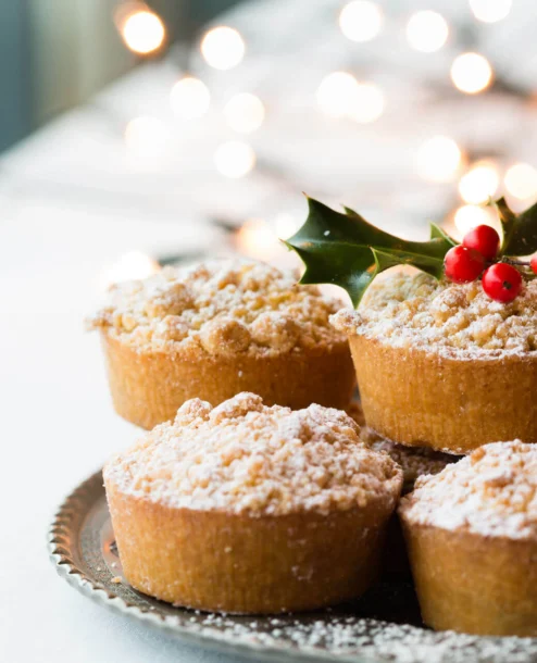 Festive mince pies with holly decoration on a plate, set against soft fairy lights.