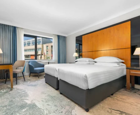 Modern hotel room with twin beds, seating area, and large window with city view.