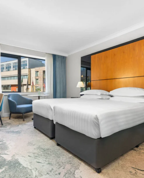 Modern hotel room with twin beds, seating area, and large window with city view.