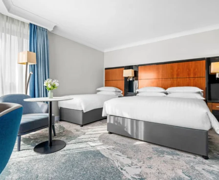 Bright hotel room with two double beds, modern decor, and a seating area by a window.