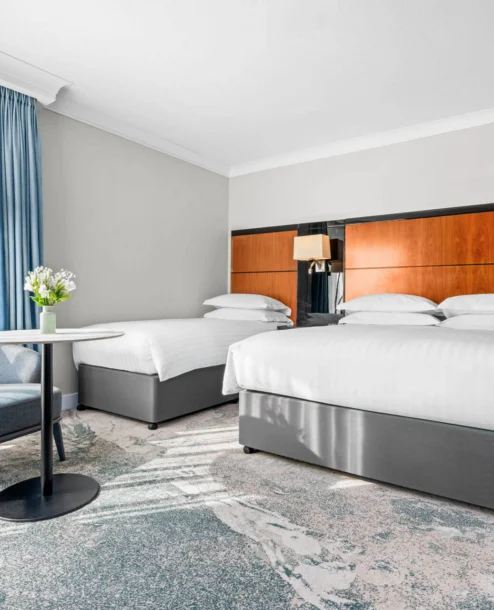 Bright hotel room with two double beds, modern decor, and a seating area by a window.