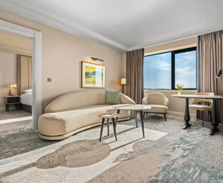 A spacious hotel suite with a cosy sitting area, large window, and a bedroom.
