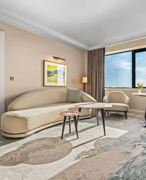A spacious hotel suite with a cosy sitting area, large window, and a bedroom.