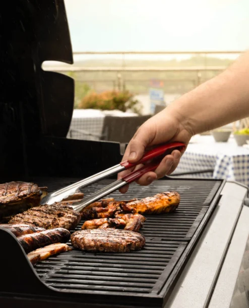 A person grills meat and corn, with tables set for dining on a sunny terrace at The Galmont Hotel.