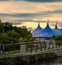 Family strolls by river near blue tent, lush greenery at sunset.