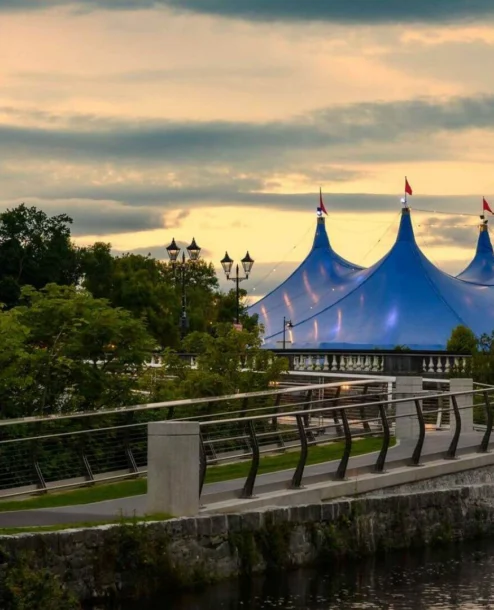 Family strolls by river near blue tent, lush greenery at sunset.