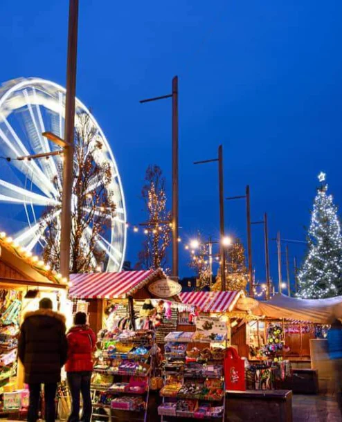 Festive Christmas market with families shopping near a lit Ferris wheel and Christmas tree.