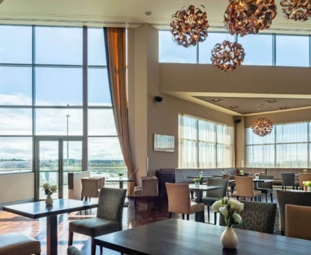 Sunlit hotel dining area with large windows, elegant lighting, and neatly set tables.