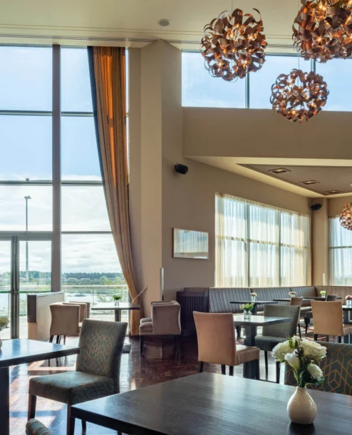 Sunlit hotel dining area with large windows, elegant lighting, and neatly set tables.