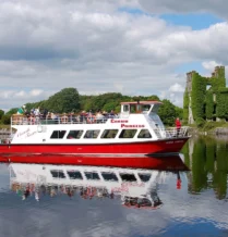 Families enjoying a scenic boat tour near a historic ivy-covered ruin on a sunny day.