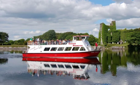 Families enjoying a scenic boat tour near a historic ivy-covered ruin on a sunny day.