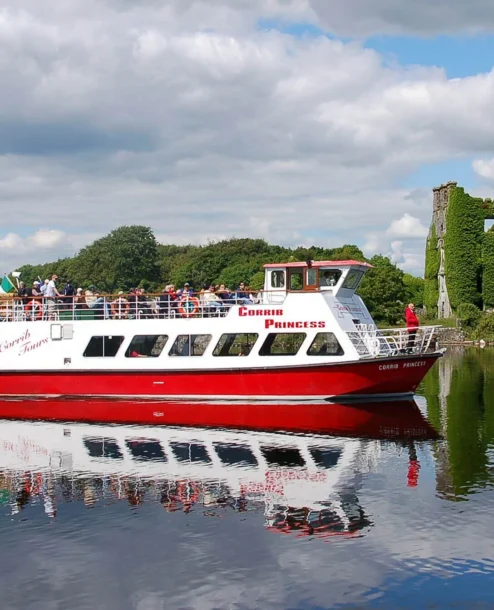 Families enjoying a scenic boat tour near a historic ivy-covered ruin on a sunny day.