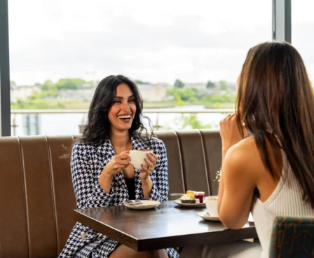 Two women enjoying coffee at The Galmont Hotel, with scenic views through large windows.