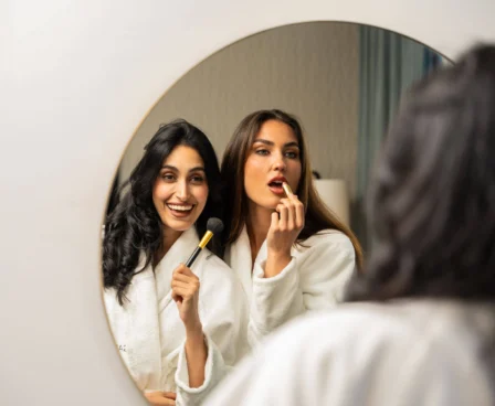 Two women in robes apply makeup cheerfully in a hotel room mirror.