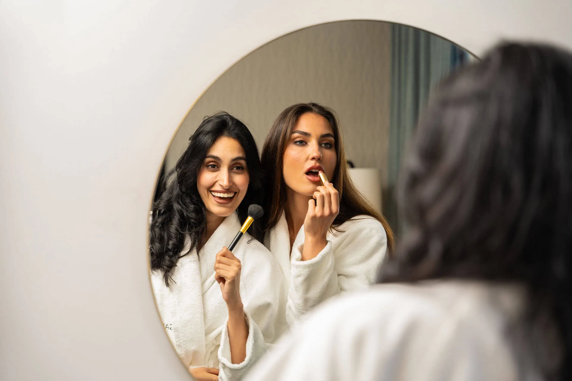 Two women in robes apply makeup cheerfully in a hotel room mirror.