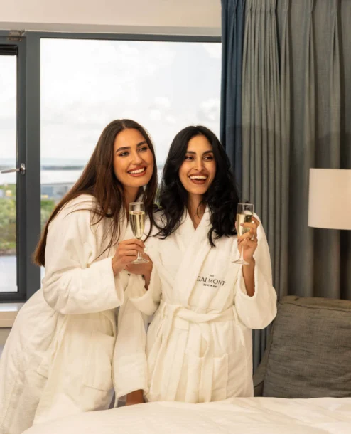 Two women in robes enjoy champagne in a cosy hotel room with a scenic view at The Galmont.