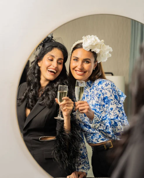 Two smiling friends toast with champagne in elegant hotel room mirror reflection.