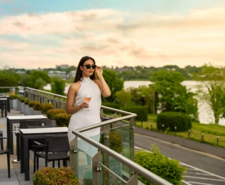 Woman enjoying a drink on a hotel balcony overlooking a scenic riverside at sunset.