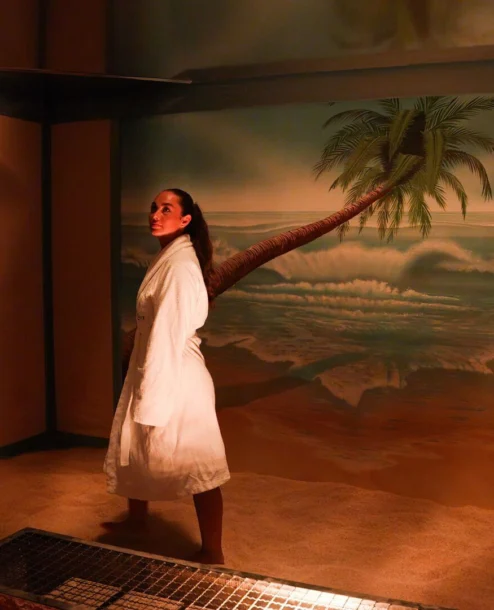 Woman in white robe stands in tropical-themed spa room at The Galmont Hotel.