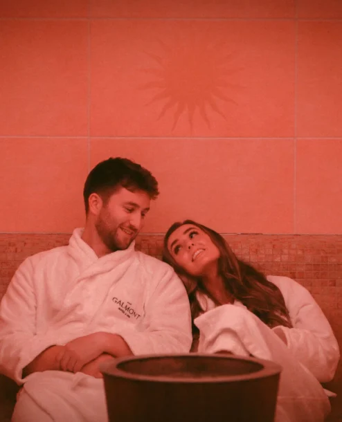 A couple relaxes in cosy robes at The Galmont Hotel & Spa sauna, with warm amber lighting.