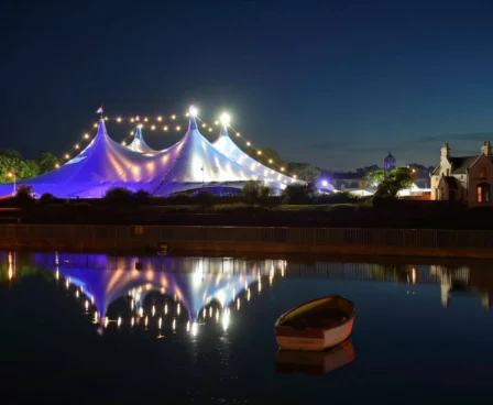 Illuminated tent by water at dusk near serene hotel setting.