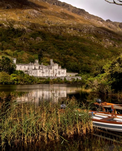 Elegant hotel by serene lake with mountains; a boat and lush greenery in the foreground.