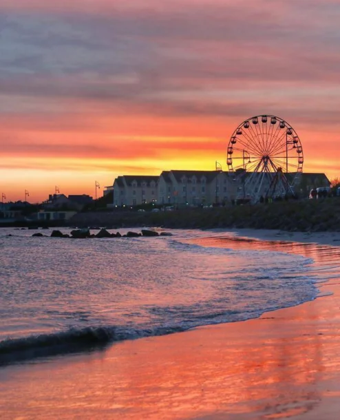 Family strolling by the beach near The Galmont Hotel & Spa at sunset with a ferris wheel in view.