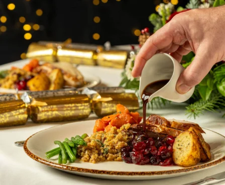 A hand pours gravy over a festive Christmas dinner with vegetables and stuffing on a decorated table.