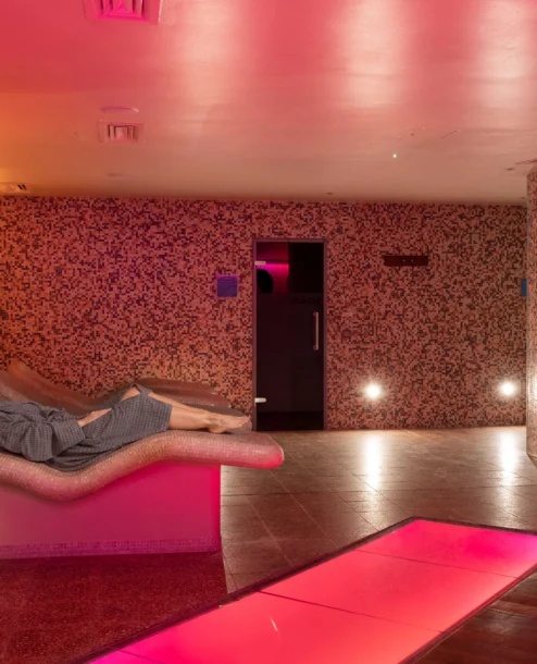 A person relaxes on a lounge chair in a dimly lit, tranquil spa room with warm lighting.