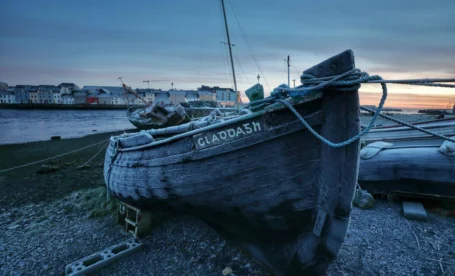 Frosted wooden fishing boat on a chilly harbour shore at sunrise, family walking by riverside town visible across water (boat name CLAODA5h).